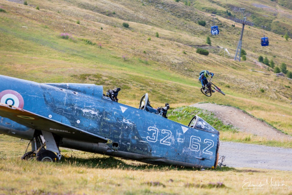 In primo piano il rottame di un aereo della seconda guerra mondiale mentre sullo sfondo Giacomo Bisi salta in mtb un panettone nel bike park del Mottolino a Livigno